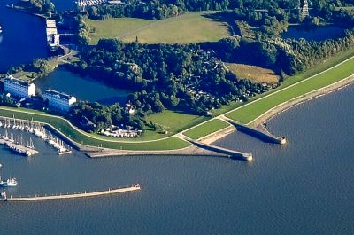 Luftbildaufnahme aus größerer Höhe. Zwischen dem Meer am unteren Bildrand und dem Innenhafen oben liegt eine Landfläche aus Grasflächen und Baumbeständen. Zwei parallele Molen ziehen sich vom Meer durch den Landbereich in den inneren Hafen.