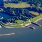 Luftbildaufnahme aus größerer Höhe. Zwischen dem Meer am unteren Bildrand und dem Innenhafen oben liegt eine Landfläche aus Grasflächen und Baumbeständen. Zwei parallele Molen ziehen sich vom Meer durch den Landbereich in den inneren Hafen.